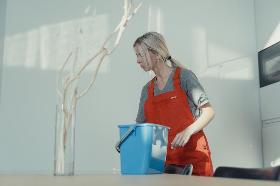 A professional cleaner, likely a woman, smiling and engaging in a surface cleaning task inside a well-lit room with natural light streaming through sheer curtains. She holds a blue cloth in her right