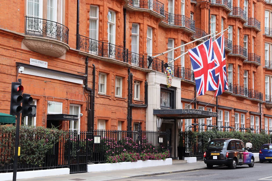 The exterior facade of The Dorchester, a luxury hotel situated in Mayfair, featuring large glass windows and elegant metal railings on the balconies. The building's beige stone walls are accented with