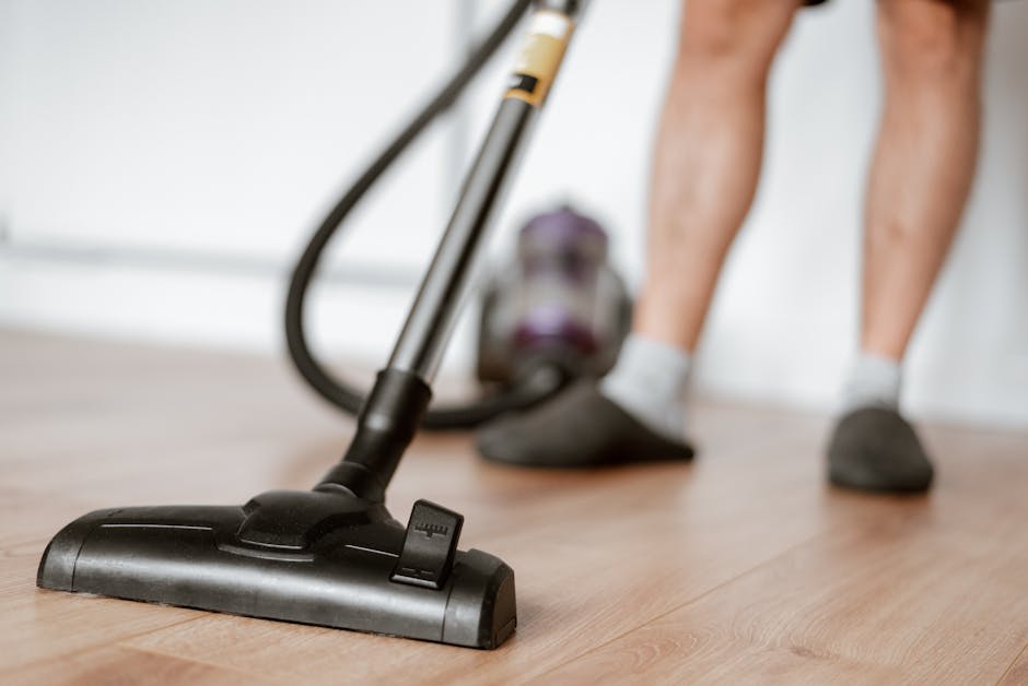 Close-up image showing a person vacuuming a wooden floor with a black and silver vacuum cleaner, focusing on the vacuum head and the person's legs dressed in grey socks and dark slippers. The scene appears well-lit, emphasizing the clean and polished surface of the floor. The vacuum cleaner's hose is connected to the main body, which is positioned behind the person, likely in a residential space. The image highlights surface cleaning and maintenance, typical of domestic cleaning tasks offered by Mayfair Cleaner, especially in the context of the Mayfair W1J Flat Cleaning Guide for Mount Street residents, with an emphasis on hygiene and thorough cleaning.