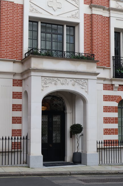 The exterior of a historic residential building in Mayfair with a brick façade accented by white stone detailing around the windows and entrance. The ground floor features a black wooden front door with decorative glass panels, framed by an elegant white stone arch adorned with intricate carvings. Above the door, there is a large window with black framing and a decorative wrought iron railing on the balcony. The entrance is flanked by a potted topiary tree and a small black metal fence runs along the sidewalk. Natural daylight illuminates the building's façade, showcasing its classic architectural style. Mayfair Cleaner’s expertise in surface cleaning and deep cleaning helps maintain the building's pristine appearance, ensuring ongoing hygiene and aesthetic appeal for residents and visitors alike.