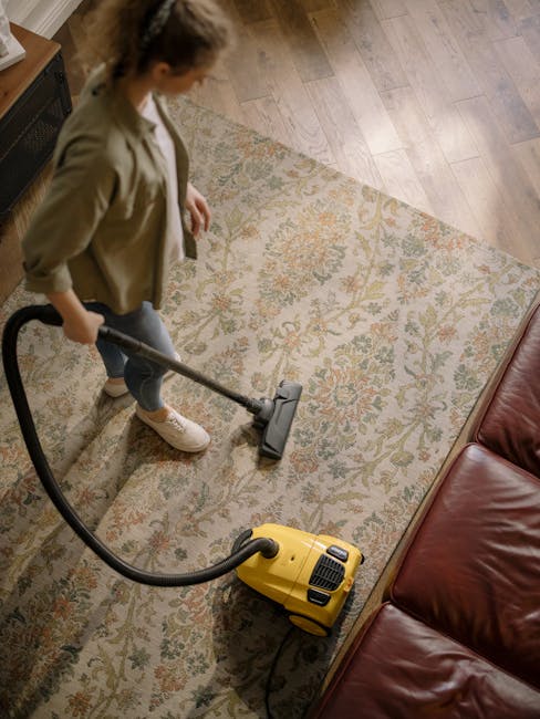 A person dressed in full protective PPE, including a white suit, gloves, and a face mask, is using a vacuum or extraction machine to deep clean a beige, plush carpet in a bright, modern living room. T